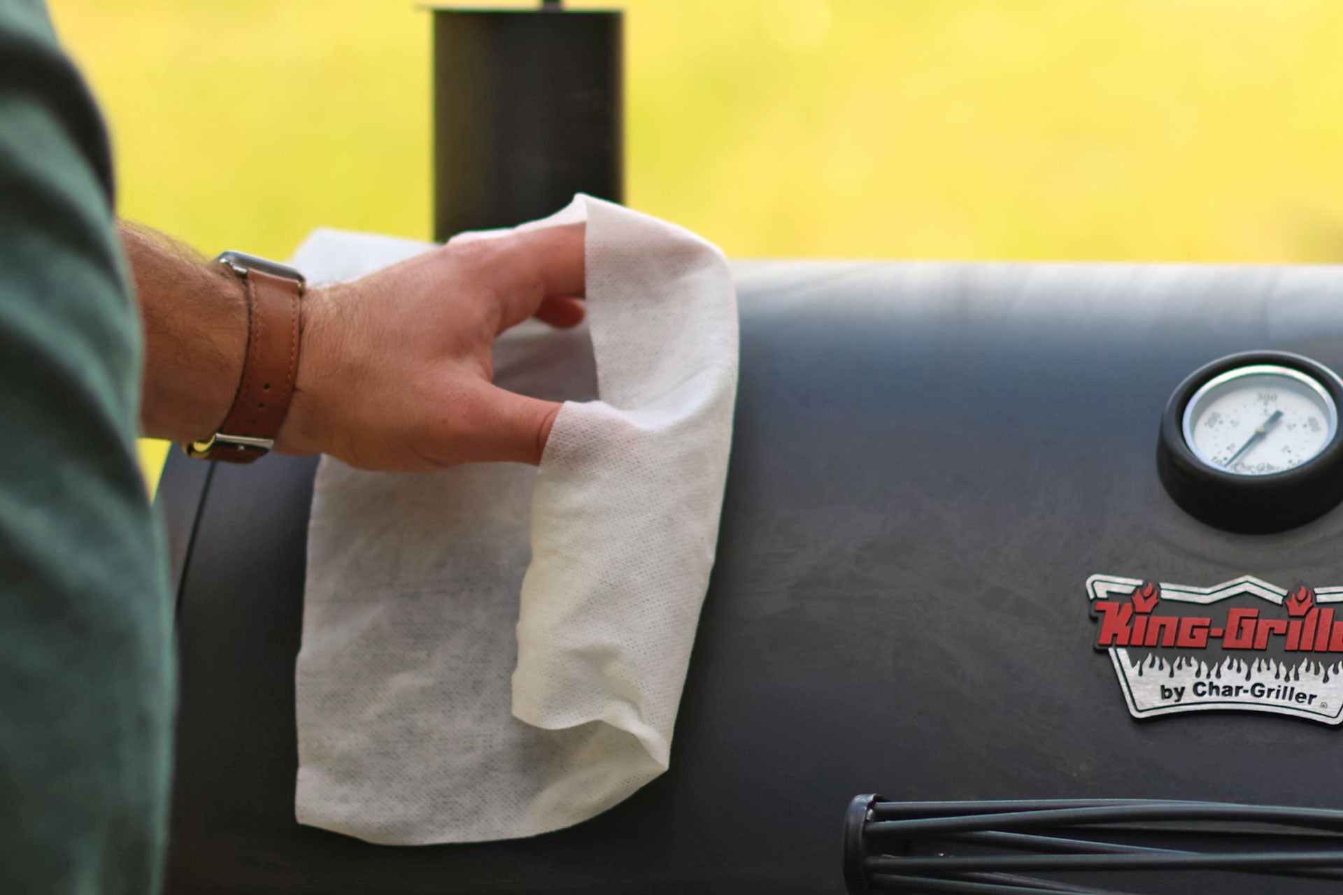 Person cleaning a grill with a cloth, with a King Grills by Char-Griller branded grill in the background.