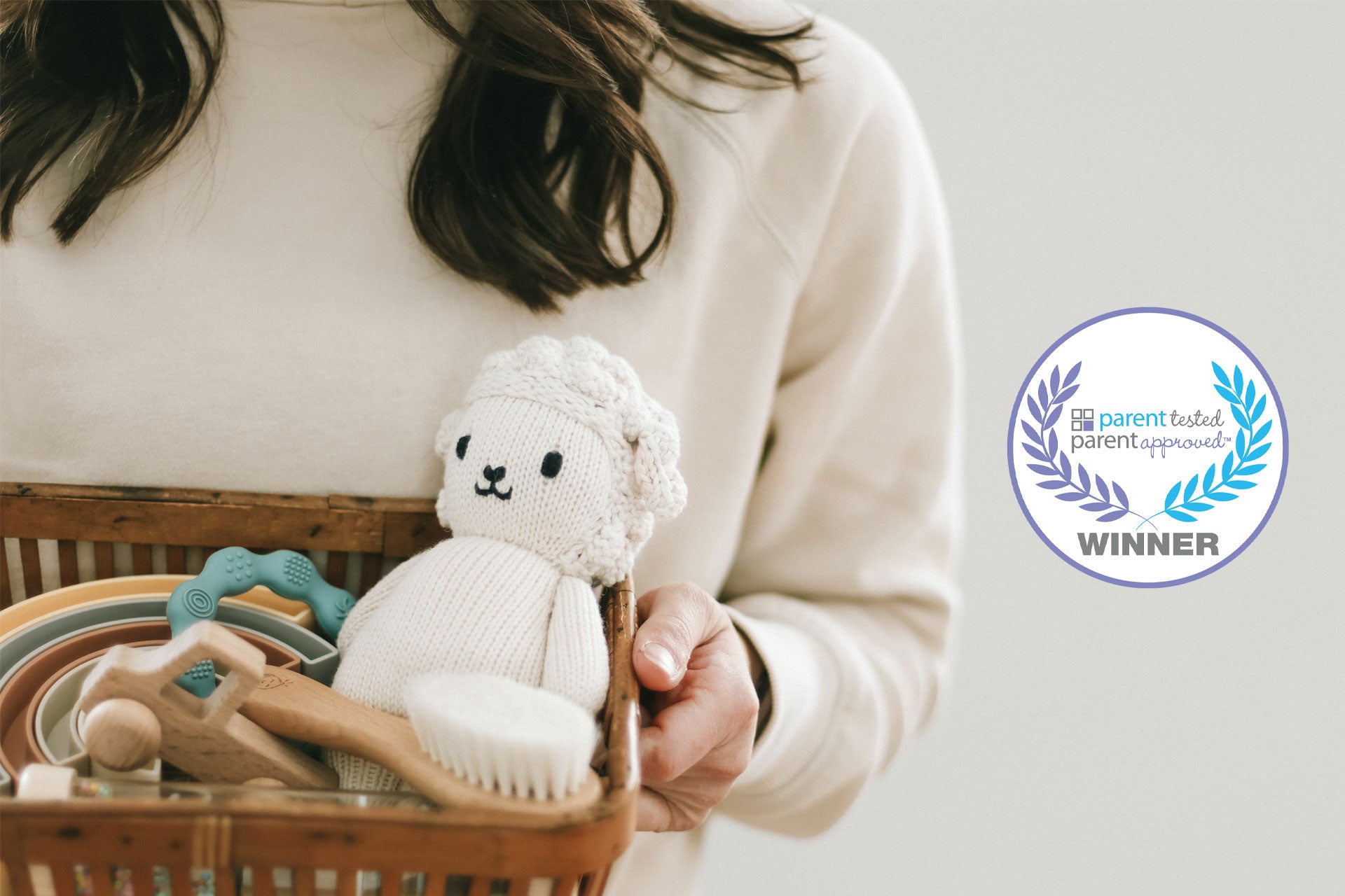 Person holding a basket with toys, including a white knitted toy, against a neutral background.